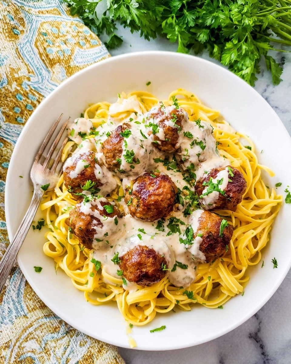 A white plate holds a serving of yellow spaghetti pasta twisted into a nest at the bottom, topped with seven round, browned meatballs scattered over the pasta. Creamy white sauce with herbs is drizzled generously over the meatballs and pasta, with small green parsley pieces sprinkled on top. A silver fork rests on the plate's edge, partially on the pasta. The dish is set on a white marbled surface with a patterned cloth and fresh green parsley in the background. Photo taken with an iphone --ar 4:5 --v 7