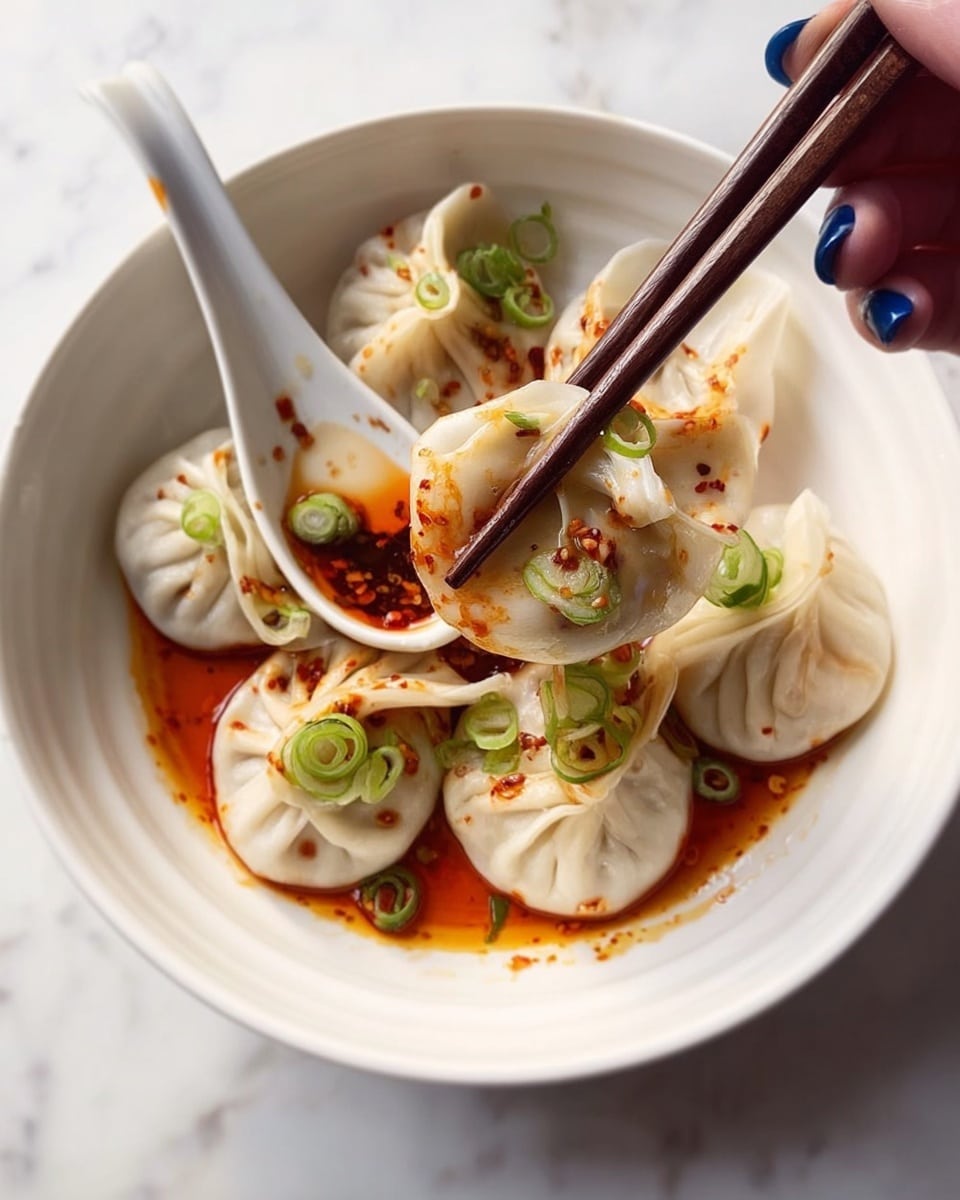 A white bowl holds several round dumplings with pleated edges, each topped with small pieces of bright green sliced scallions and a drizzle of reddish-brown spicy sauce that pools lightly at the bowl's base. A pair of dark brown chopsticks lifts one dumpling, showing translucent, slightly crinkled skin, partly dipped in the sauce held by a white ceramic spoon. A woman's hand with blue nail polish gently holds the chopsticks over the bowl. The background is a white marbled surface. Photo taken with an iphone --ar 4:5 --v 7