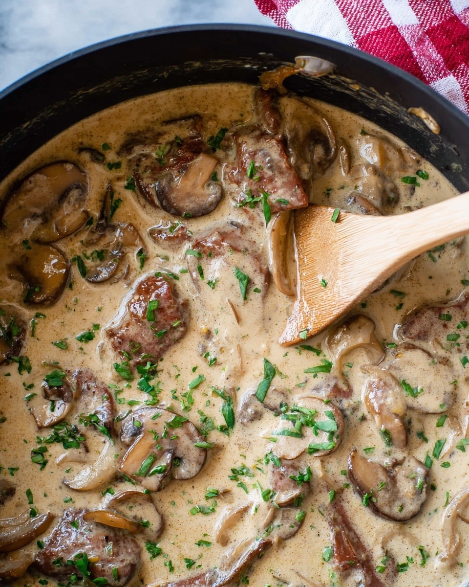 The image shows a creamy dish in a dark pan with several small slices of brown meat and light brown mushroom slices covered in a thick beige sauce. Small bright green chopped herbs are scattered evenly on top. A smooth light wooden spoon is partially dipped into the sauce on the right side. The background is a white marbled surface with a red and white checkered cloth partially visible. photo taken with an iphone --ar 4:5 --v 7